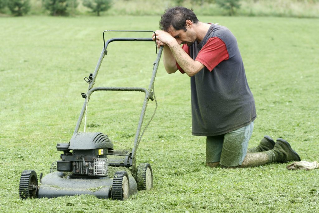 A man on his knees with his lawn mower, tired from mowing his lawn