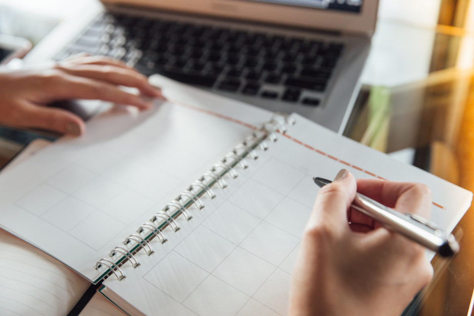 Woman in casual dress sitting at table in cafe and writing in calendar notebook.