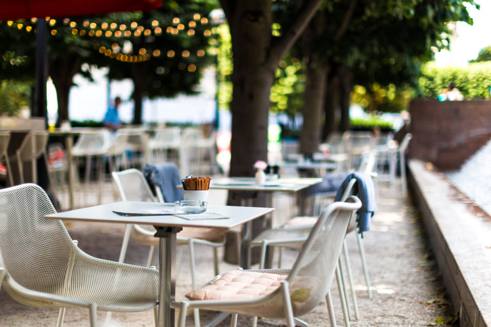 Color image depicting chairs and tables arranged at an outdoors cafe in central London, UK. It is a beautiful green space lined with trees and illuminated with fairy lights. There are bowls of sugar on the tables, but there are no people around.