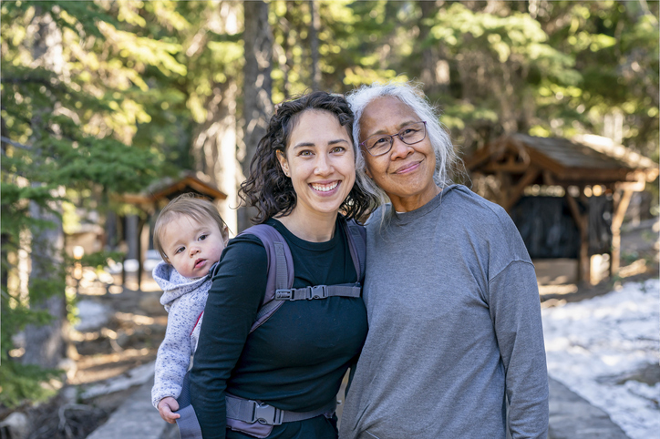 A young mixed race woman carries her one year old daughter on her back in an ergonomic baby carrier while out on a hike with her senior mother. The multi-generation family is staying active and healthy outdoors in Oregon.
